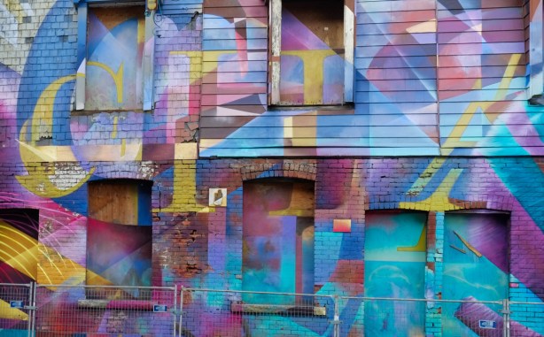 part of a street art mural with the word change written in gold and yellow on a multi coloured background, on old houses with boarded up windows