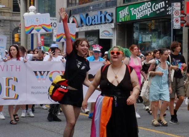two happy women marching in dyke parade for pride 2023 in Toronto, one has green hair and a rainbow flag tucked into the waistband of her skirt