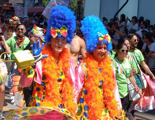 two people walking in pride parade, large blue fuzzy hair wigs, orange boas, rainbow bows in the hair