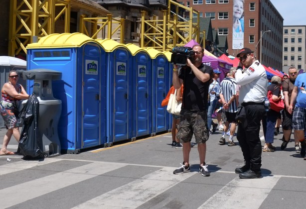 a security guard stands beside a man using a large video camera, along side a row of porta potties