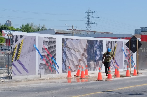 a workman in a blue hardhat cleans the street between hoardings with large photos on them and orange cones blocking traffic