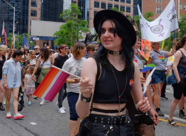 dyke march, pride toronto, woman in black hat, short black top and carrying pink orange and white striped flag