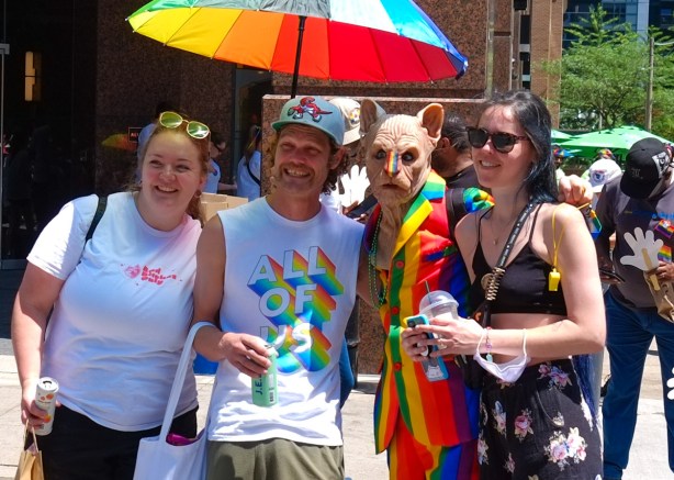 four people posing under a rainbow umbrella inclusing one person in an animal mask