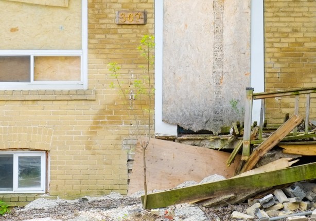 boarded up doors and window of an empty brick house, wood porch has rotted away