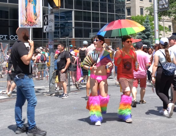 two men and a rainbow umbrella, one in pink bikini bottoms and both in fuzzy rainbow coloured leg warmers