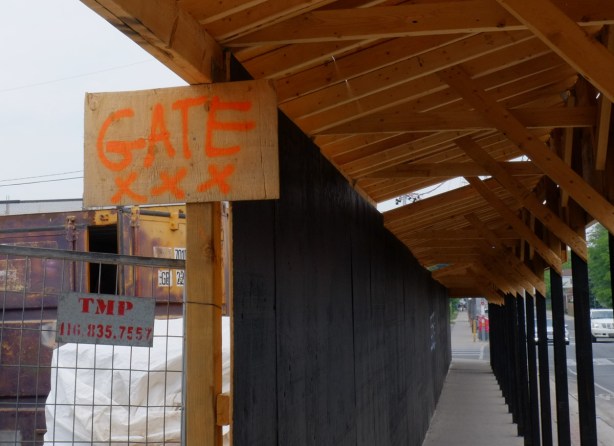 at an entrance gate to construction site, beside a covered sidewalk, hand written sign says gate xxx