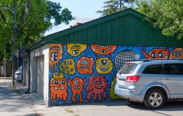 grey car parked beside a garage with street art mural painted on it. Lots of red, orange, and yellow, blob-like characters