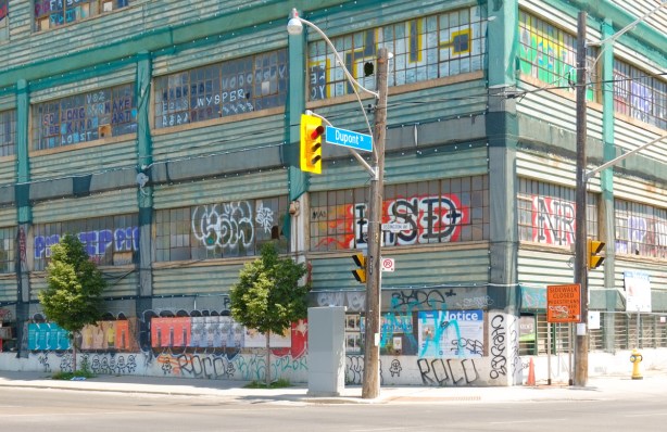 old industrial warehouse building on the northeast corner of Dupont and Ossington, teal cloured cladding with lots of small square window panes, graffiti tags all over the windows, posters along the wall at street level 