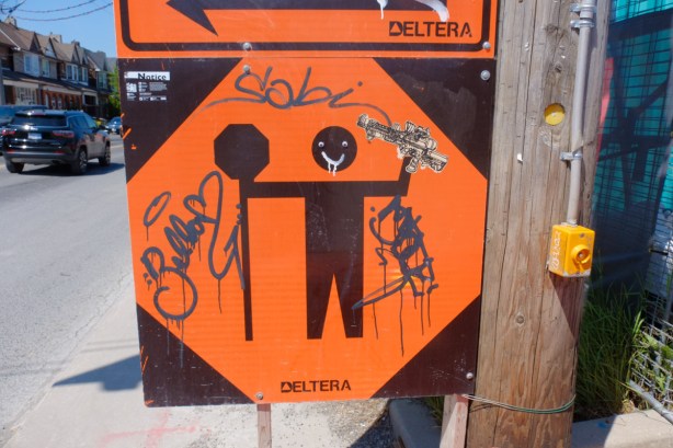 black and orange construction sign with figure of man holding sign, happy face added to head and a machine gun put in his hands, altered sign