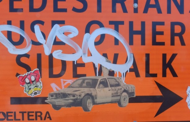 orange construction sign, pedestrians use other sidewalk, with stickers and pasteups on it, an old car in black and white