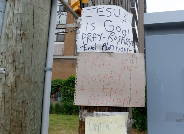 hand written signs and posters on a utility pole, two of them are pray the rosary to end abortion, jesus is god