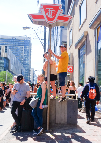 people watching pride parade and Yonge and Dundas, standing up on railing around subway entrance