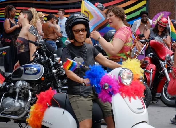 people walking in dyke March, woman in black helmet sitting on white scooter