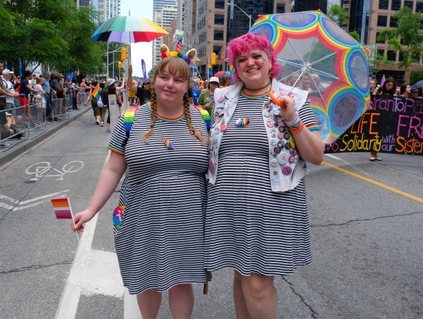 two woman on Bloor street, pride weekend, in matching blue and white small striped dresses, one holding a clear umbrellas with rainbow decoration