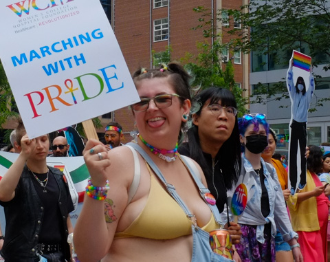 women in dyke march along Bloor street in Toronto, woman in yellow bra holding a sign, sticking her tongue out at camera