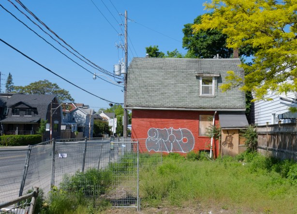 vacant building beside a grassy space, red brick, tagged on the side, old grey shingle roof, boarded up lower level window, construction fence around it 
