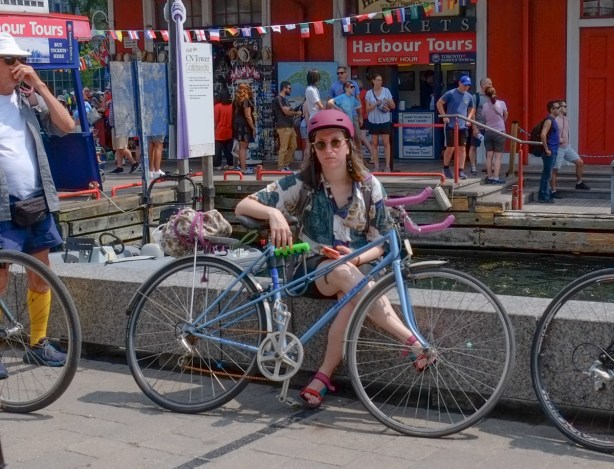 a bored looking woman witting on a concrete barrier, holding her bike in front of her, by the waterfront