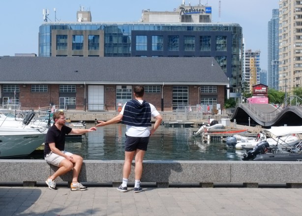 two young men sitting by waterfront, each reaches his hand towards the other