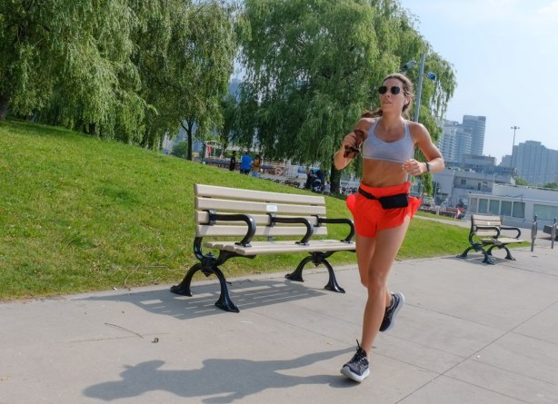 a woman in bright orange shorts jogs along the waterfront in front of a green park and wood bench