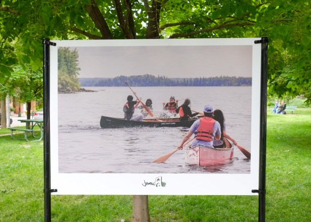 colour photo of people in two canoes, paddling on a lake, on display in a park, 