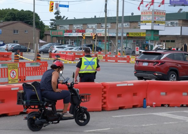 a man on a motorbike rides past orange barricades at construction site, policeman patrolling the traffic at the intersection, Keele and Finch, LRT construction, stores on other corner of intersection in the background