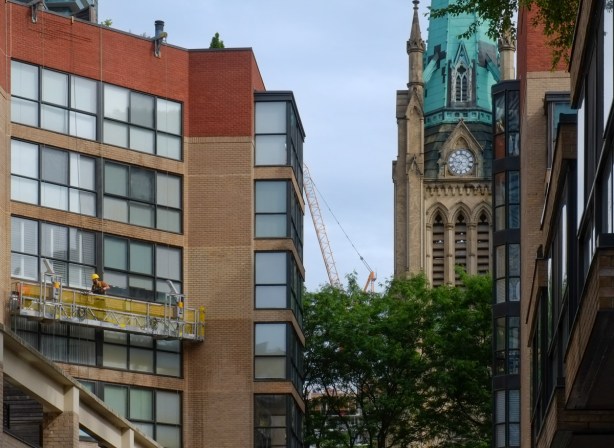 The green weathered copper roof of St. James Cathedral steeple can be seen in the gap between two newer low rise buildings, one of which has a platform of window cleaners on it 