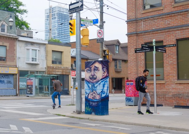 at Queen East and Sumach, a sidewalk box painted by Elicser Elliott of a man in a blue shirt, short dark hair. A man walks by, another man is sorting out his scooter while waiting for a traffic light