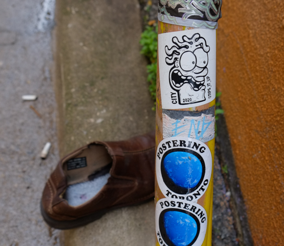 a brown shoe on the ground, stickers on a metal pipe, some say postering, another is a black drawing of a face