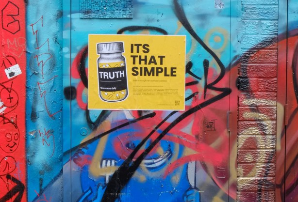 a poster pasted on top of graffiti covered wall, picture of a bottle with yellow pills and label that says truth. words beside bottle say it's that simple