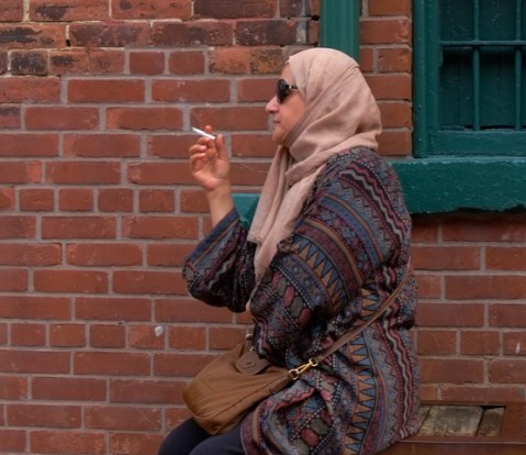 woman in pink head scarf and dark sunglasses, sitting outside brick building at distillery district, smoking a cigaretts