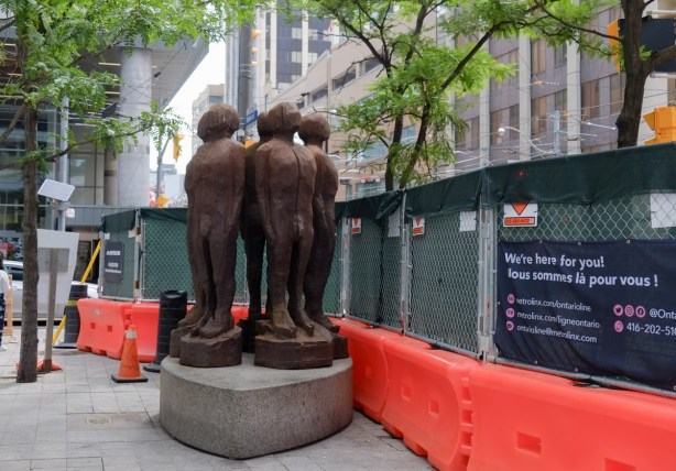 sculpture of men in circle with backs together, at Church and Queen, now beside fence and hoardings for Queen street closure
