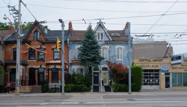 two houses in a row, one is light blue with a large rose bush in front, to the right is a mechanic shop, Central Auto 