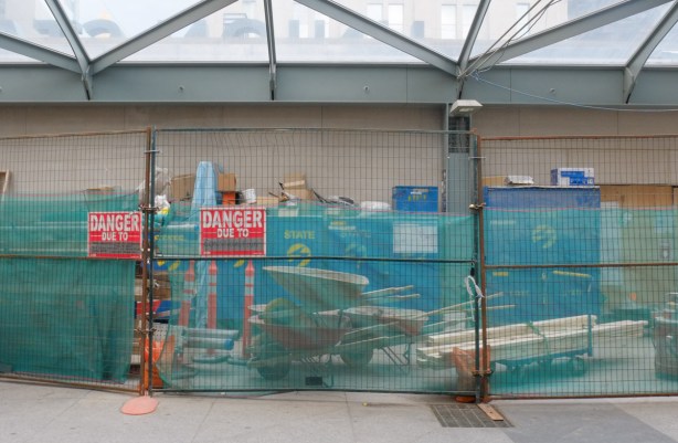 construction and maintenence equipment like a wheelbarrow kept behind a green mesh fence, with danger due to construction signs on it 