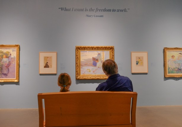 a father and daughter witting on a bench looking at paintings in an art gallery