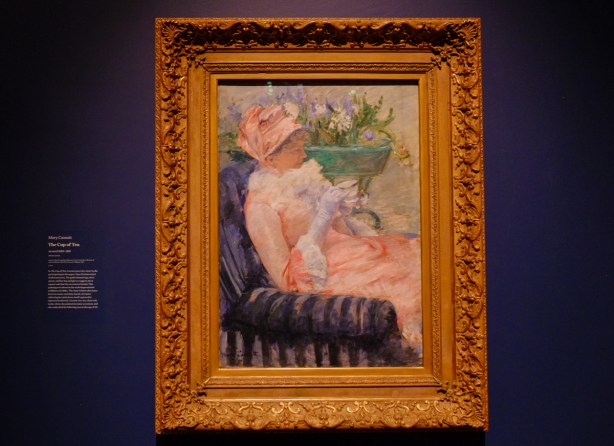 painting on a wall in a gallery, woman all dressed up in pink with a cup of tea in her hand