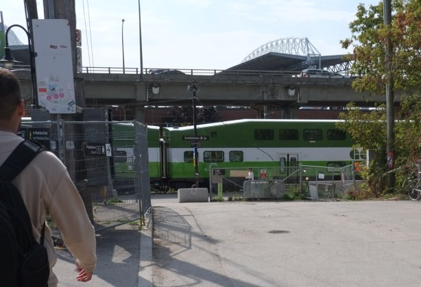 a man with a backpack walks towards Exhibition station where there is a green and white GO train already in the station