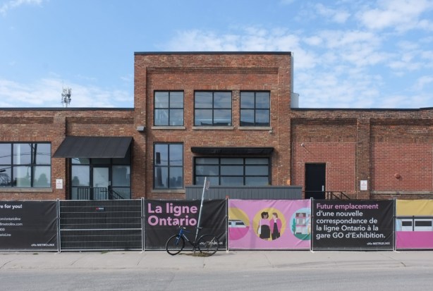 low rise older brick building, light industrial, with black hoardings in front, with signs advertising metrolinx ontario line, 