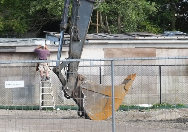 empty bucket of a front end loader sits on the ground beside a man on a ladder who is inspecting roof of building
