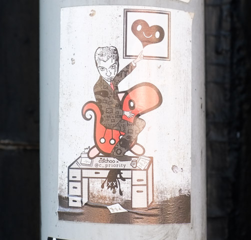 sticker on a pole, catchoo along with at priority, man on red inflatable on top of desk, pointing to picture of heart framed and hanging on a wall
