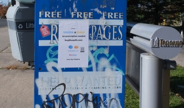 signs on the side of a blue and white free newspaper box, one sign says help wanted, and the other is an ad for viagra and cialis
