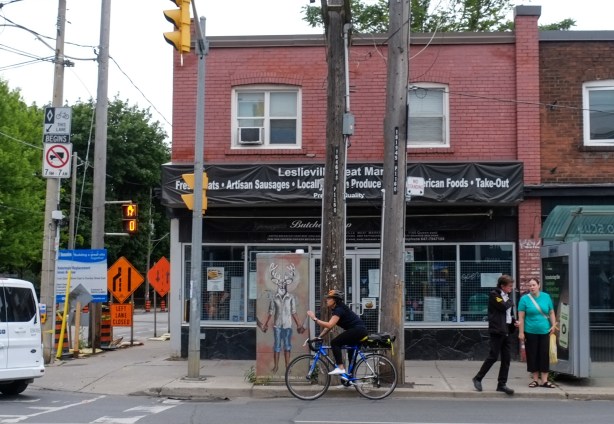 Leslieville meat market, with street art image on door, a cyclist in front, women waiting at bus stop