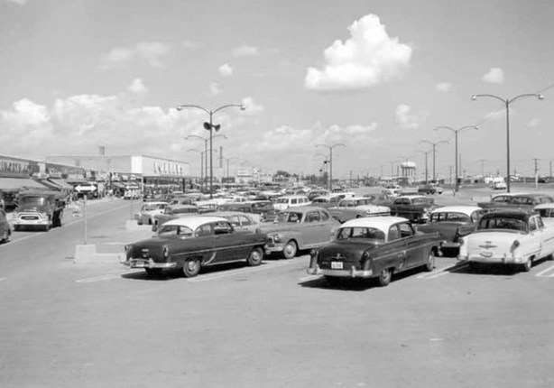 black and white photo from 1950s of cars in a parking lot in front of a mall, the golden mile mall