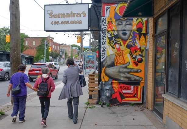 people walking past Samairas, with a street art painting on one of the exterior walls, large hand