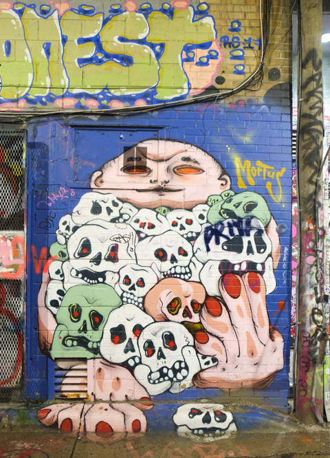 blob like figure with bald head and big hands, holding a lot of skulls, mural in graffiti alley by mortus 