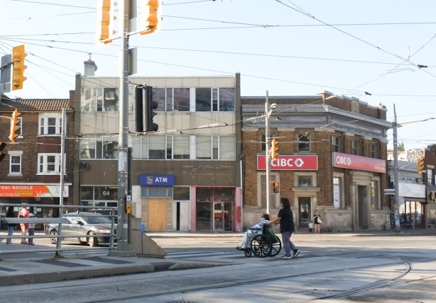 person in wheelchair being pushed across St. Clair at Vaughan Road, southwest corner with CIBC bank in old brick building