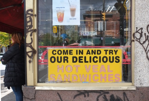 a woman stands beside a store, in window of store is a yellow sign that says come in and try our delicious hot veal sandwiches