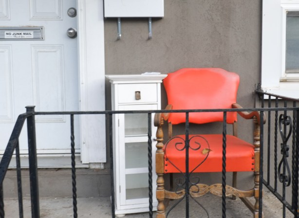 an orange vinyl padded chair on a porch with black wrought iron railing and a small white bookcase
