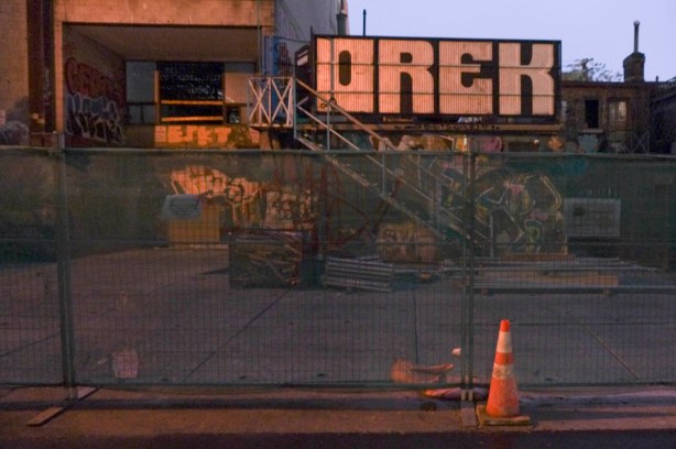 back of buildings on Queen West, now dark, with construction fence, view from graffiti alley, large orek tag on building, dark, no lights,