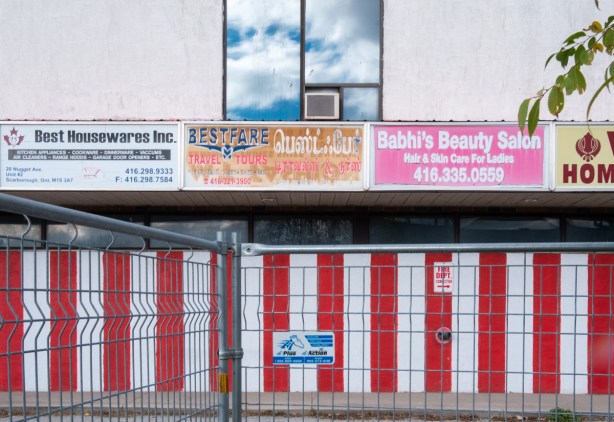 empty building behind construction fence, red and white vertical stripes on lower level, window on upper level, three signs for businesses, Best Housewares Inc., BestFare travel and tours, and Babhis Beauty Salon