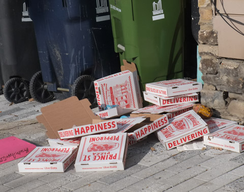 a pile of empty pizza boxes on the sidewalk beside some city of toronto rubbish bins and recycling bins